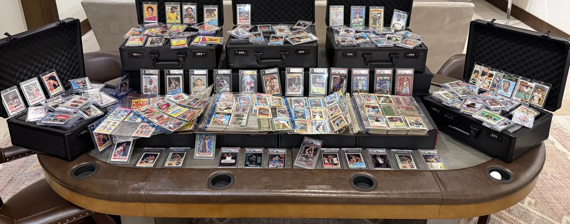 The founder&rsquo;s collection laid out on a card table: open carrying cases of vintage basketball and baseball cards in top-loaders, a row of graded cards standing along the middle, and binders of nine-pocket pages open in front.